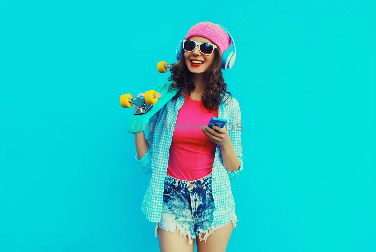 Portrait of stylish modern happy young woman listening to music in headphones with mobile phone and skateboard posing wearing colorful clothes, pink hat, sunglasses on blue studio background