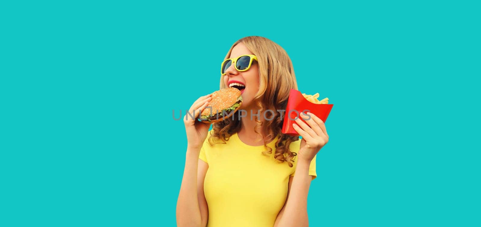 Portrait of happy cheerful young woman eating burger fast food and french fries, fried potatoes isolated on blue studio background