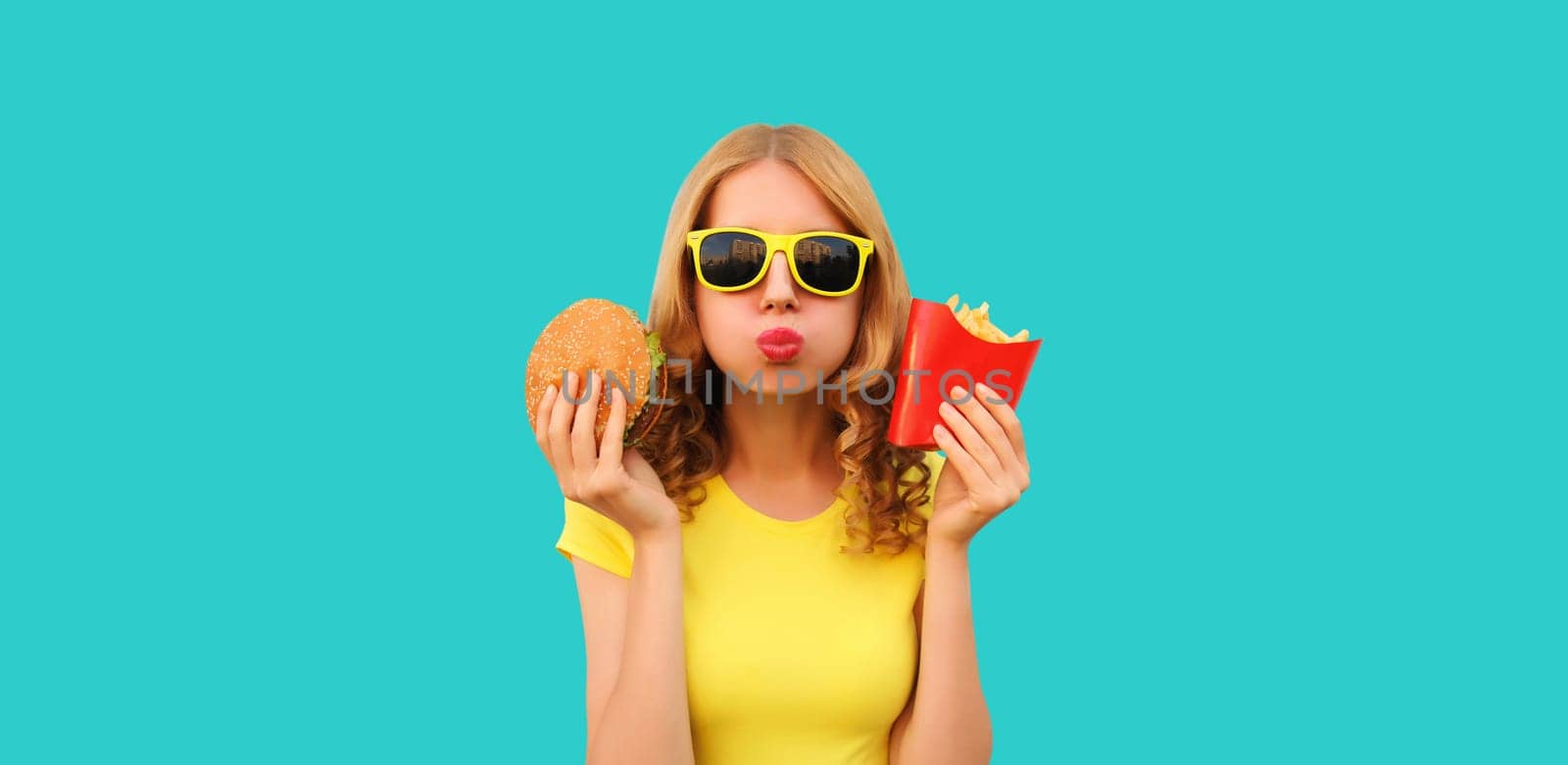 Portrait of happy cheerful young woman eating burger fast food and french fries, fried potatoes isolated on blue studio background