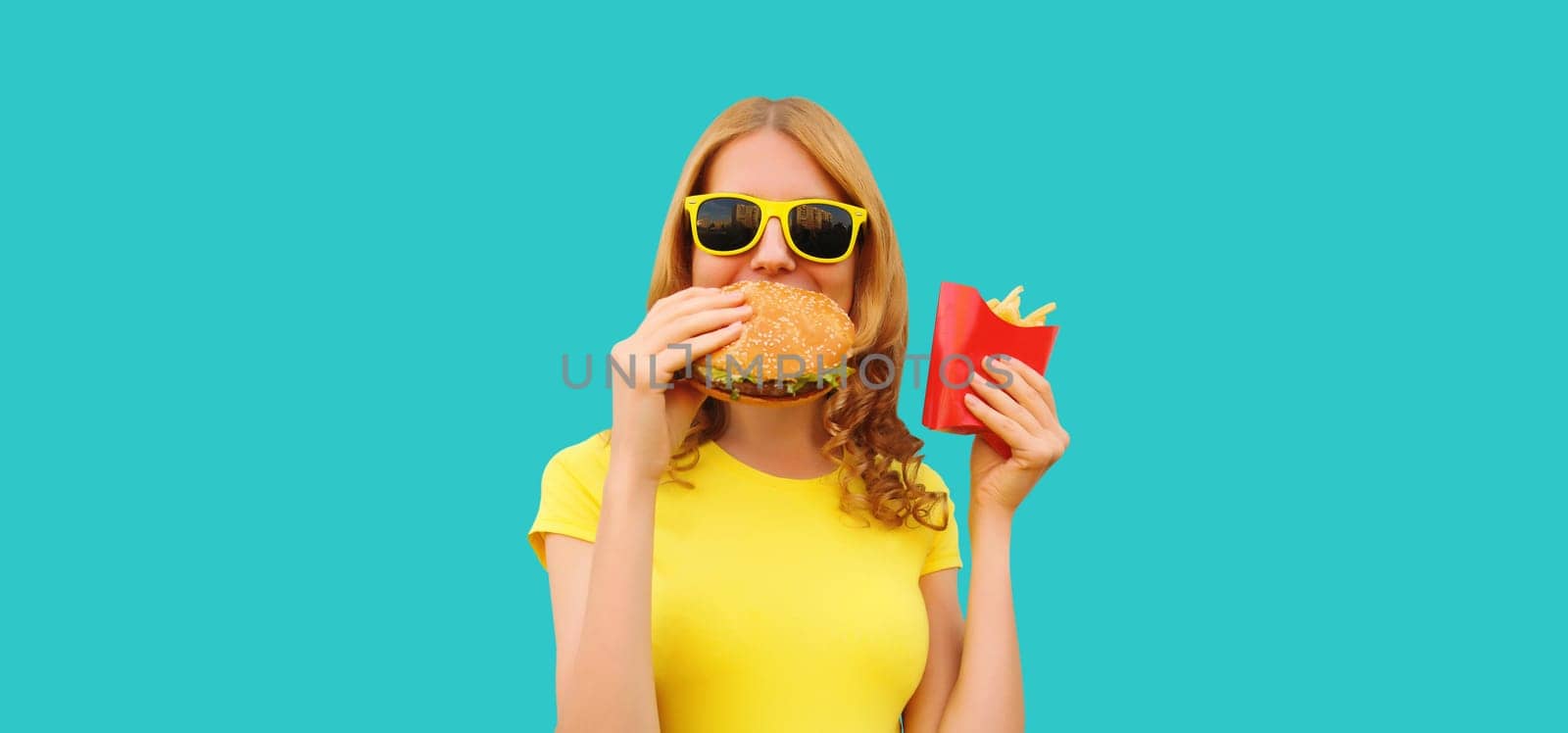 Portrait of happy cheerful young woman eating burger fast food and french fries, fried potatoes isolated on blue studio background