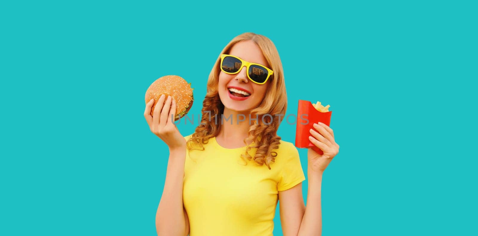 Portrait of happy cheerful young woman eating burger fast food and french fries, fried potatoes isolated on blue studio background