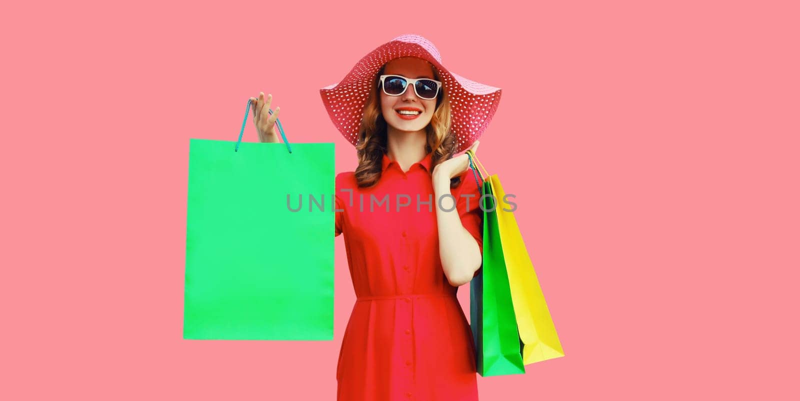 Portrait of beautiful happy smiling young woman model posing with colorful shopping bags in summer straw hat, dress on pink studio background