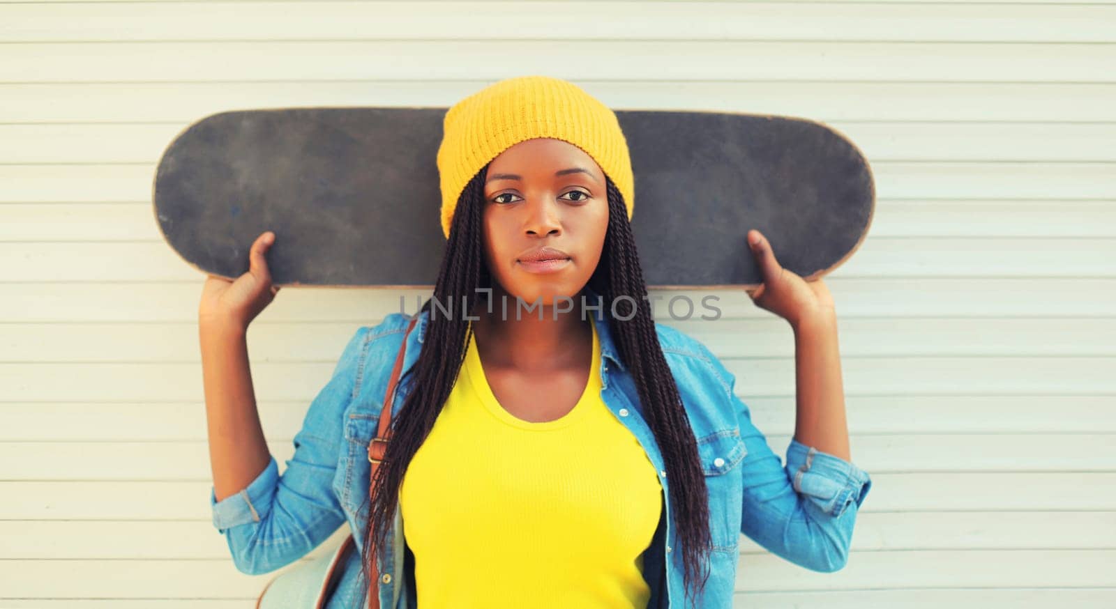 Portrait of modern young african woman model posing in colorful clothes, yellow hat with skateboard in the city