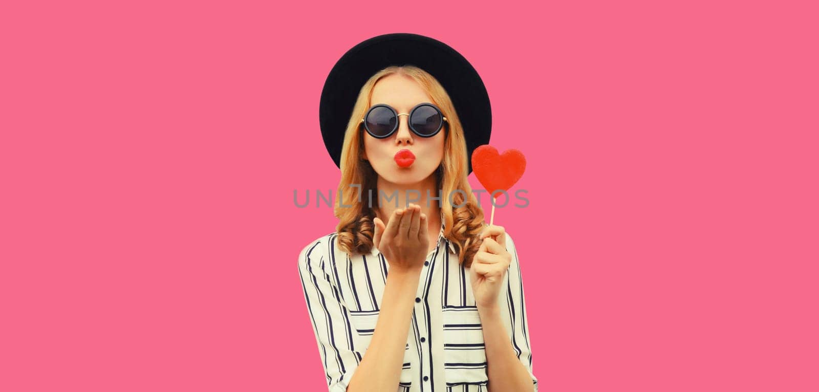 Portrait of beautiful young woman blowing her lips sends kiss with sweet red heart shaped lollipop by Rohappy