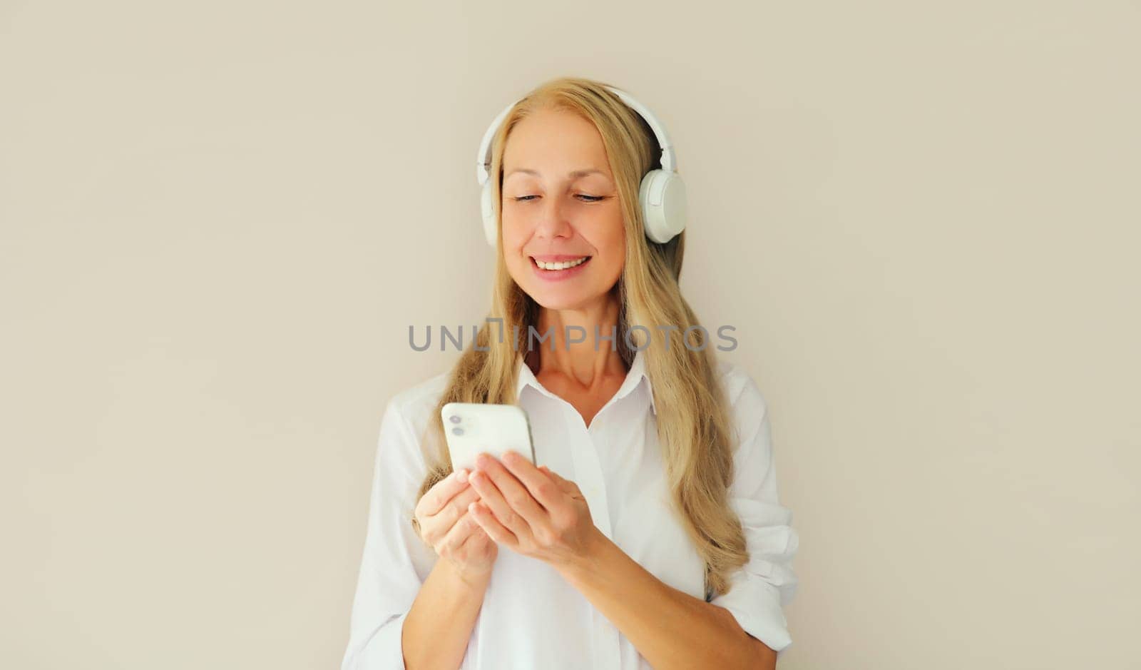 Happy smiling middle aged woman listening to music with mobile phone in headphones on studio background