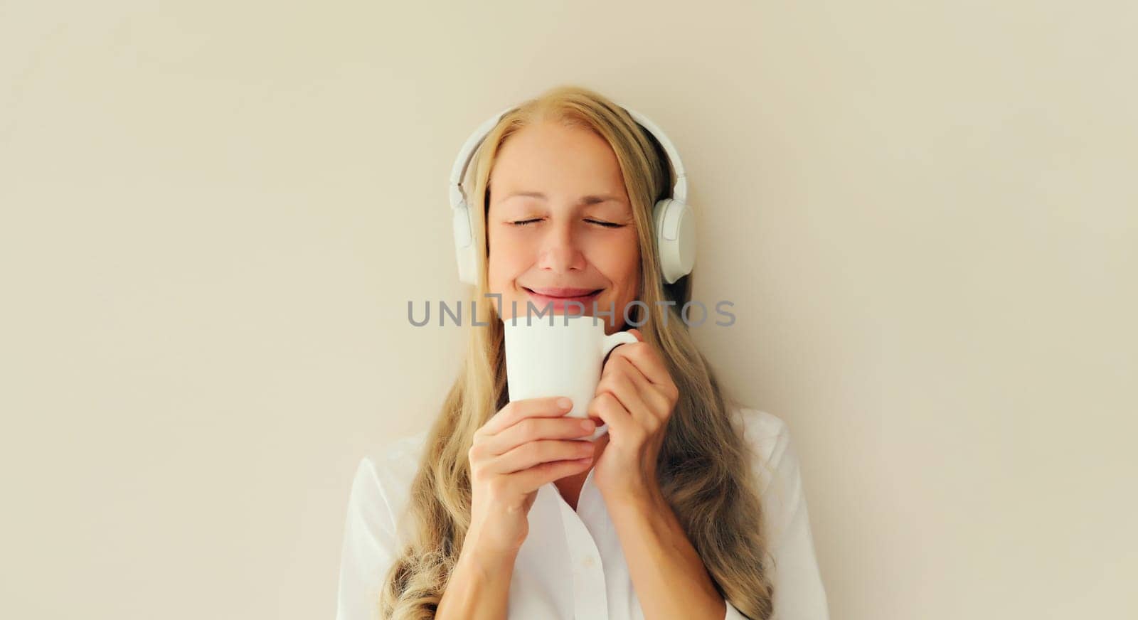 Portrait of happy relaxed middle aged woman listening to music drinking mug of coffee in headphones on white background