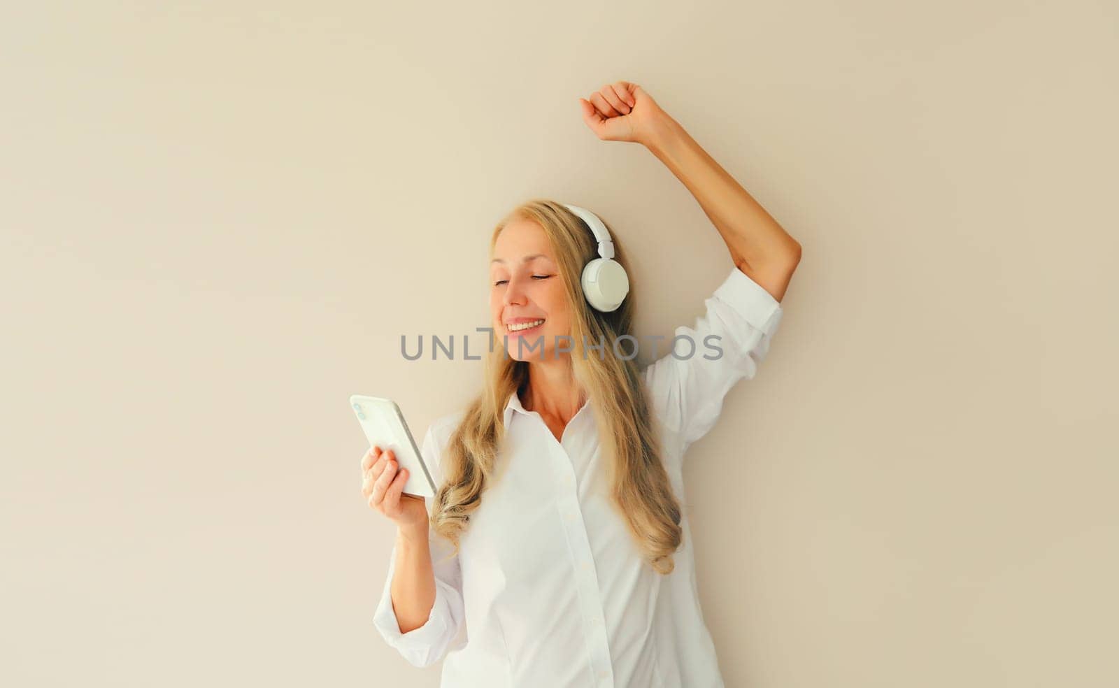 Happy cheerful middle aged woman listening to music with mobile phone in headphones having fun dancing on studio background