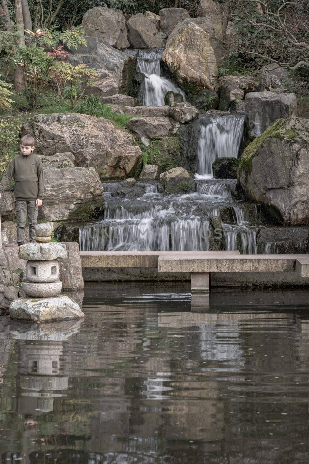 London, UK - Feb 15, 2024 - A boy stands on a stone bridge in front of waterfall and rocky cliffs in Kyoto garden, Space for text, Selective focus.
