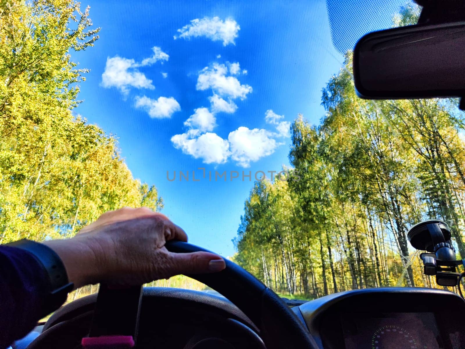 view from a car windshield of natural landscape with road, green trees and blue sky in summer or spring time. Hand of woman on steering wheel. Female traveler driving alone on trip or journey