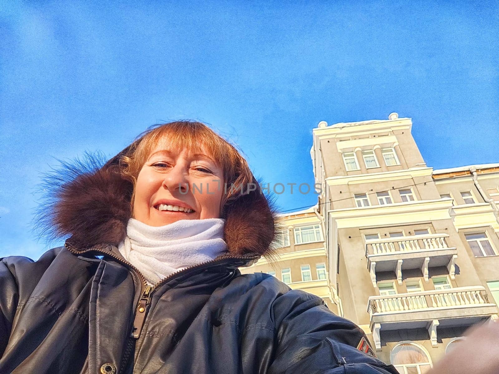 A cheerful woman poses taking selfie in front of an elegant, multi-storied building under a clear blue sky in winter, autumn or spring