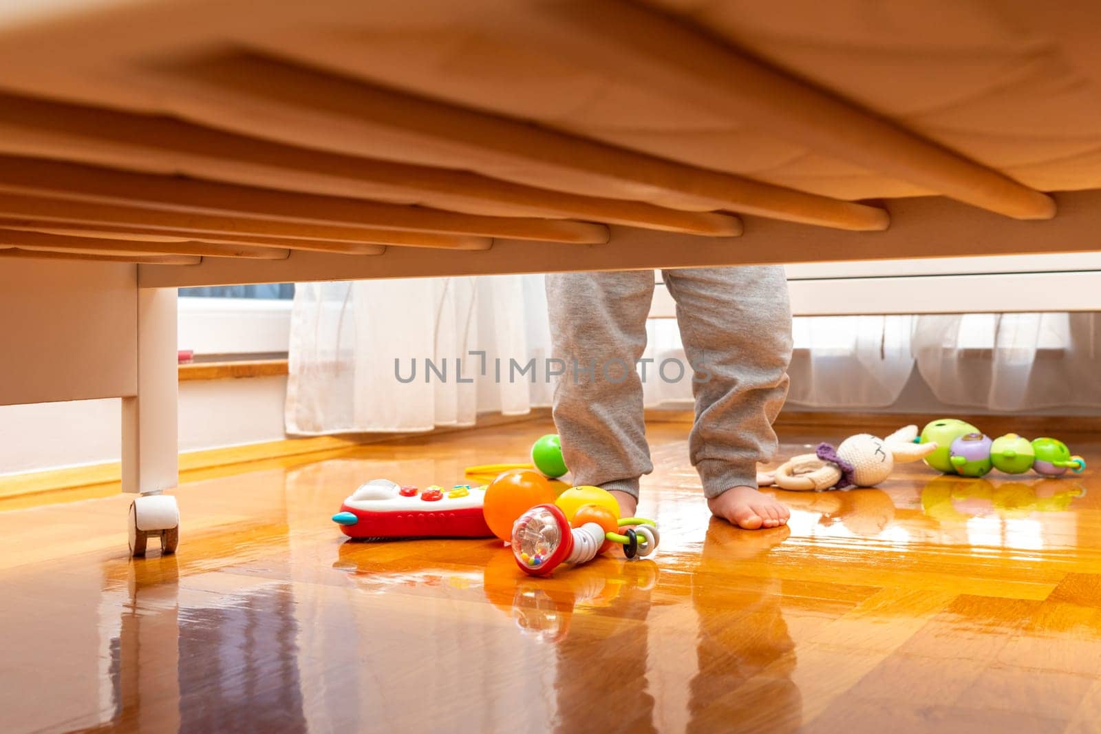 Baby standing near a crib with toys on the floor. View from under the bed. The window brings in
