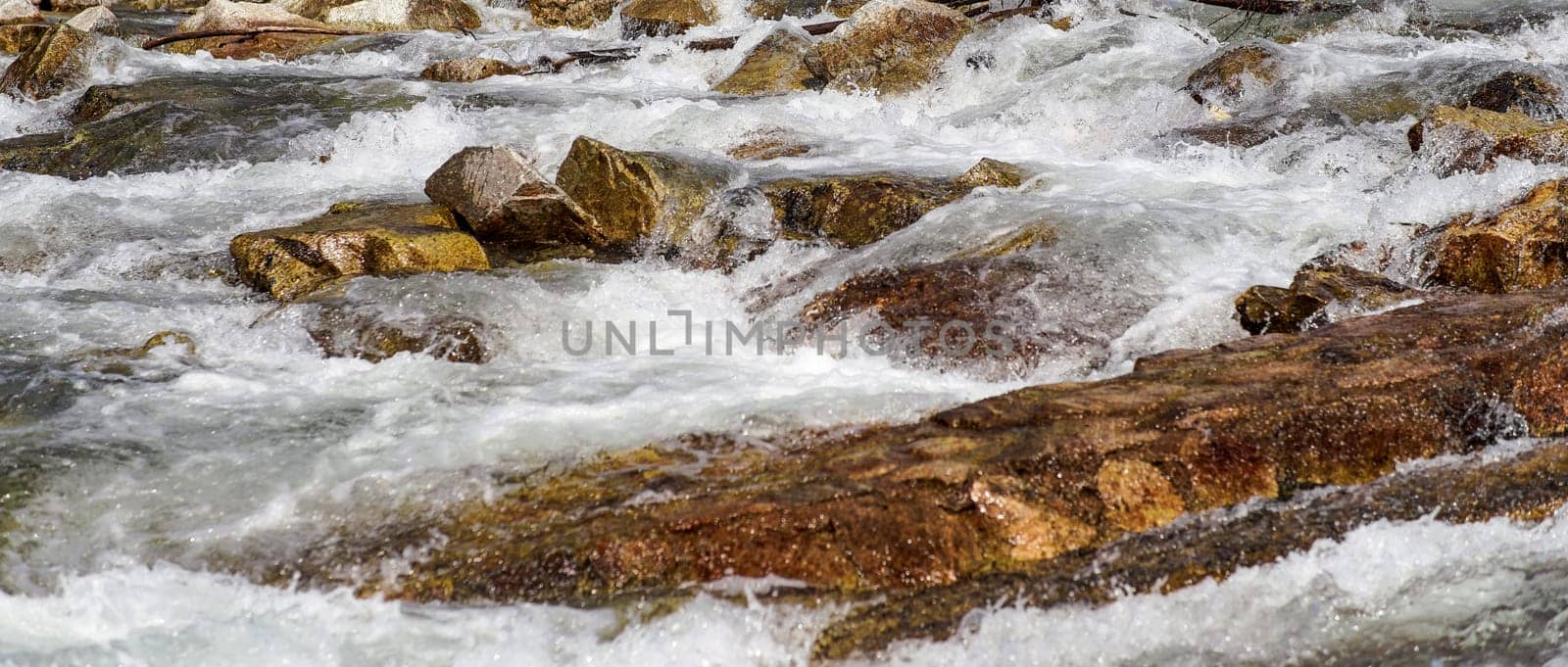 Sun shines on rapid spring river flowing over brown rocks, wide banner by Ivanko