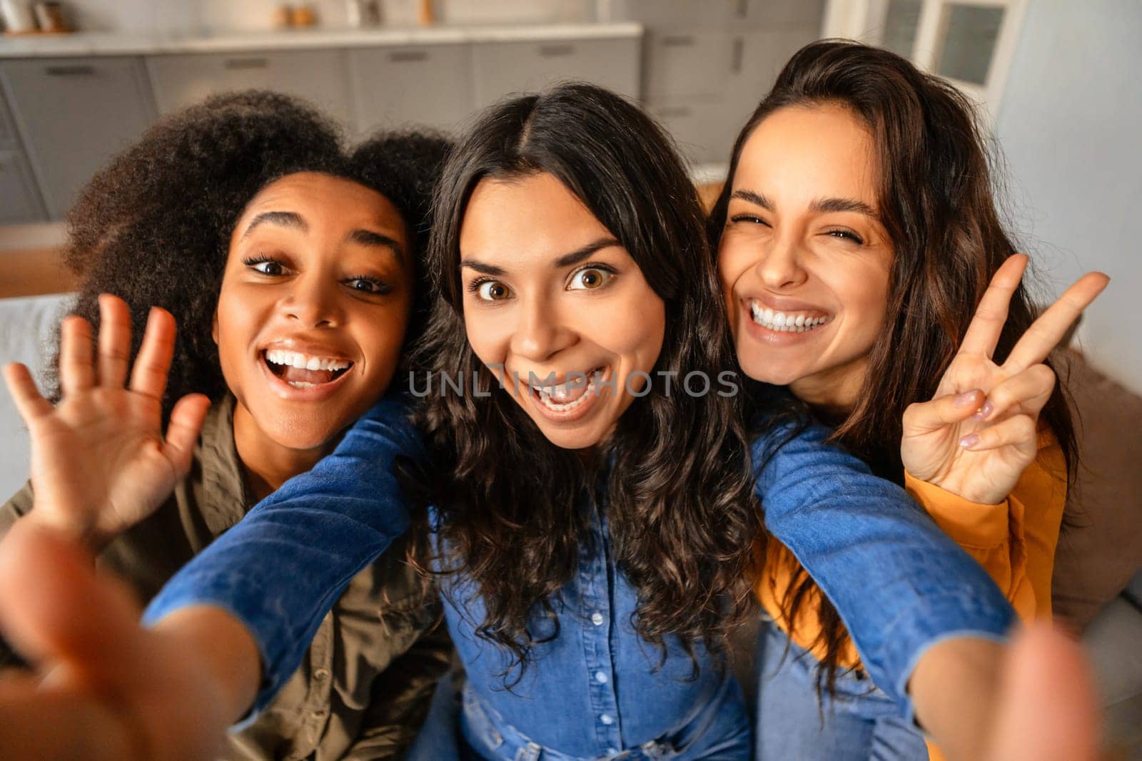 Three joyful young multicultural women taking playful selfie, waving and showing peace signs, as they posing together, making social media content at home. Diverse beauty and friendship