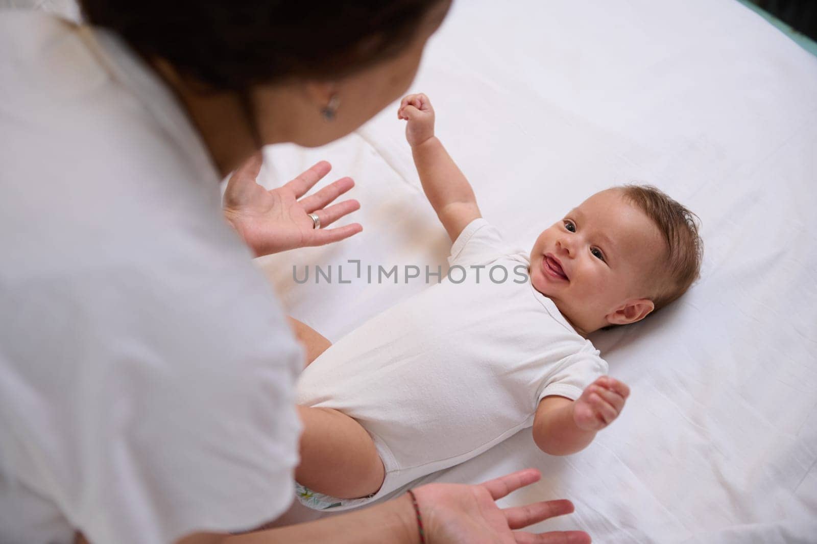 Adorable Caucasian baby boy 4 months old lying on bed, keeping his arms outstretched, smiling to his mother. View from above. Infancy. Babyhood. Maternity leave concept. Happy family relationships.