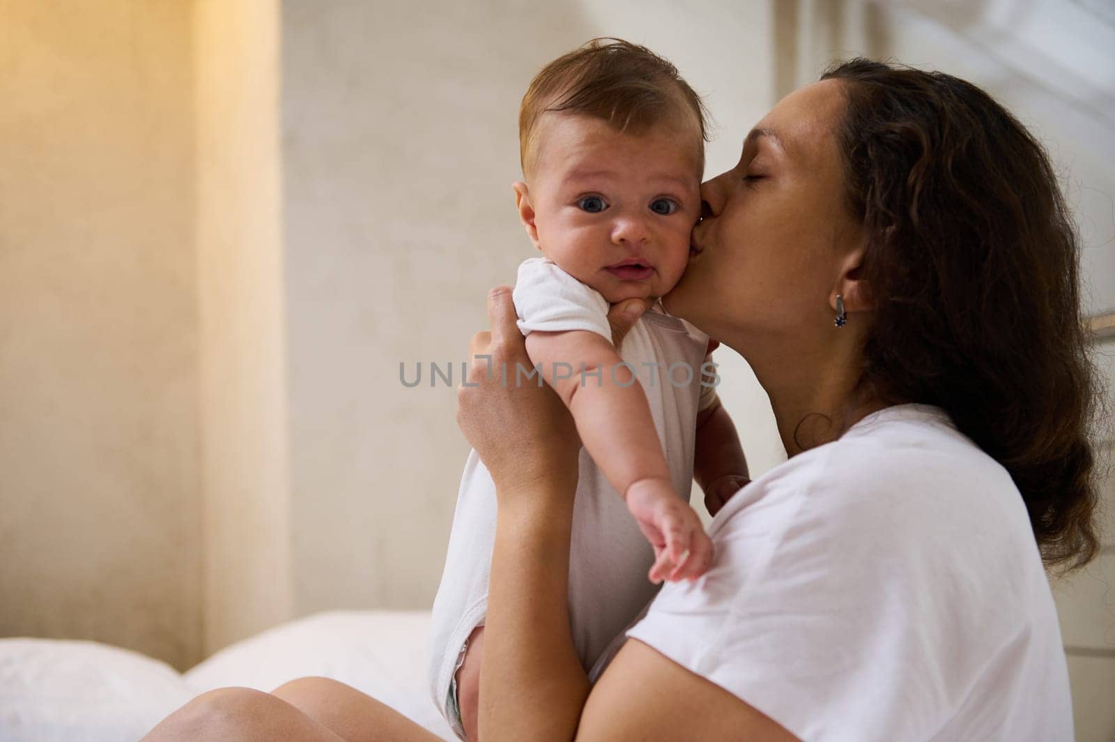 Authentic emotional portrait of a Caucasian cute baby boy, child 4 months old looking at camera while his mother cuddling and kissing him, enjoying happy time together. Babyhood. Baby care concept