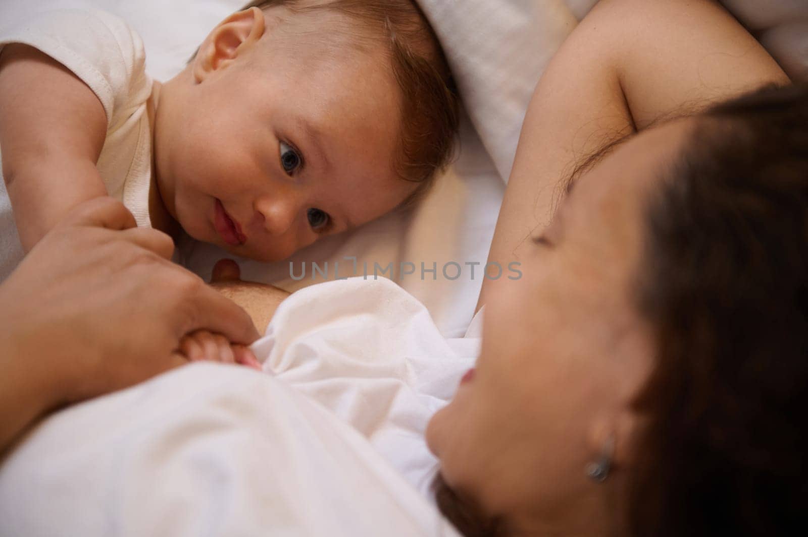 Close-up of a cute baby establishing visual and emotional contact, lying next to his mother while she breastfeeding him. Babyhood and infancy. Maternity leave. Co-sleeping and breastfeeding concept