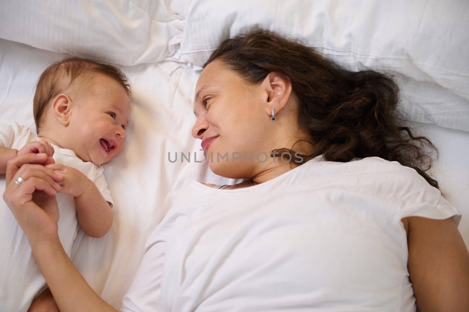 Delightful young mother lying on the bed with her adorable baby boy 4 months old. Mom and child communicating, smiling. The concept of happy maternity leave, babyhood, family relationships