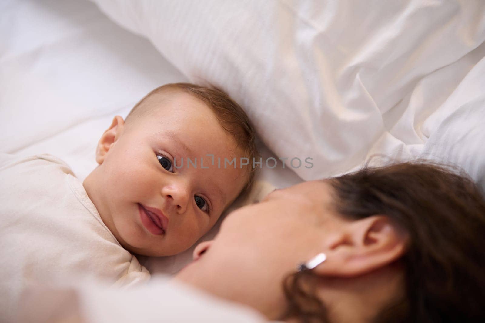 Close-up portrait of a cute baby boy looking at camera, lying on the soft white bed sheets close to his loving mother by artgf