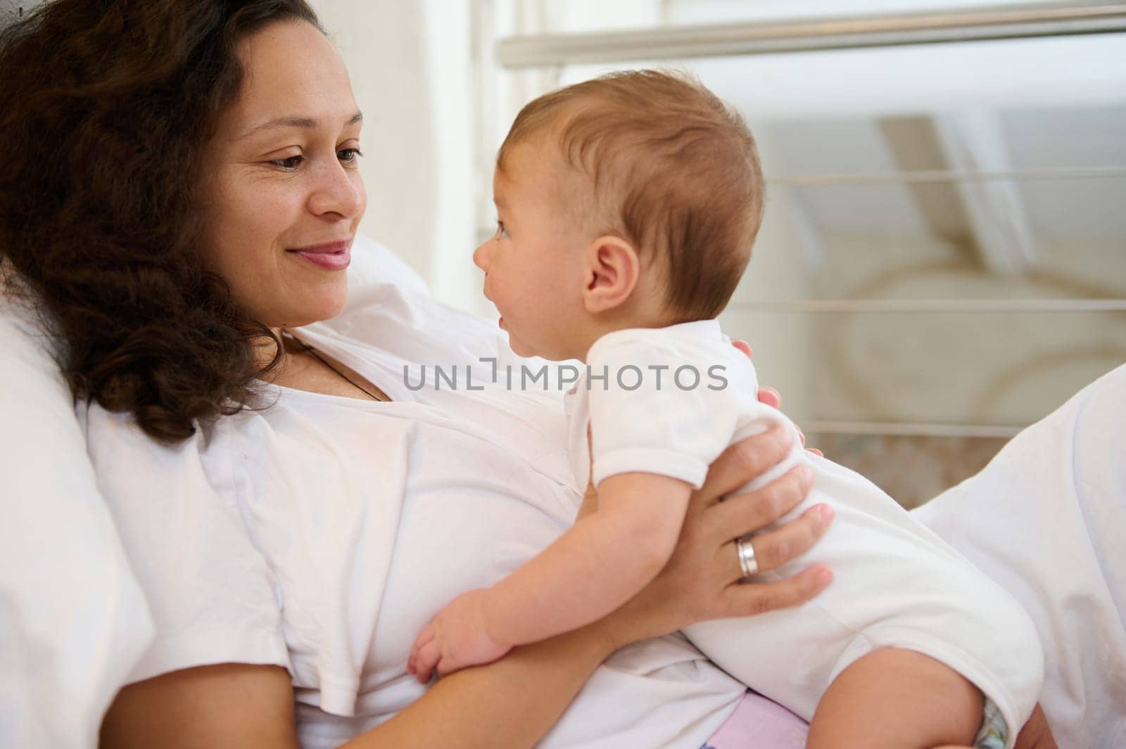 Newborn baby in tender embrace of mother. Happy delightful young smiling woman, affectionate loving mother caring of her newborn baby at home, lying together on the bed at cozy and light home interior