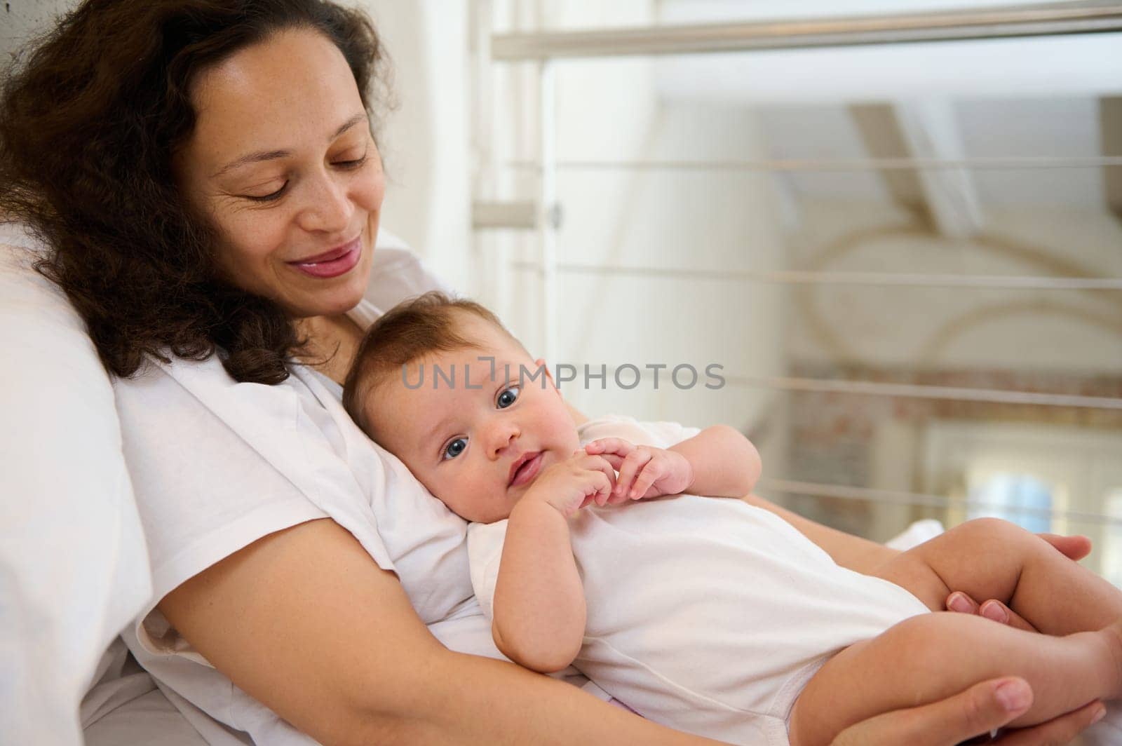 Happy young mother holding her baby while lying together on the bed at light home interior. Adorable child of 4 months old smiling cutely looking at the camera. Mother and son together. Happy family.