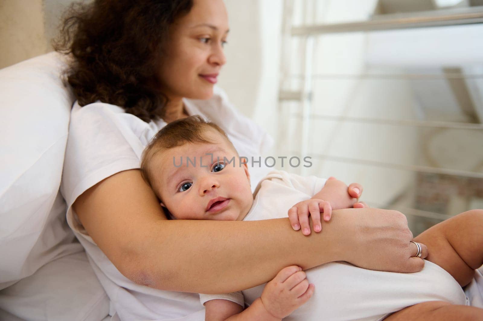 Authentic portrait of lovely baby boy 4 months old in the arms of her loving delightful mother dreamily looking away, enjoying happy time with her newborn child at home. Maternity leave. Infancy time