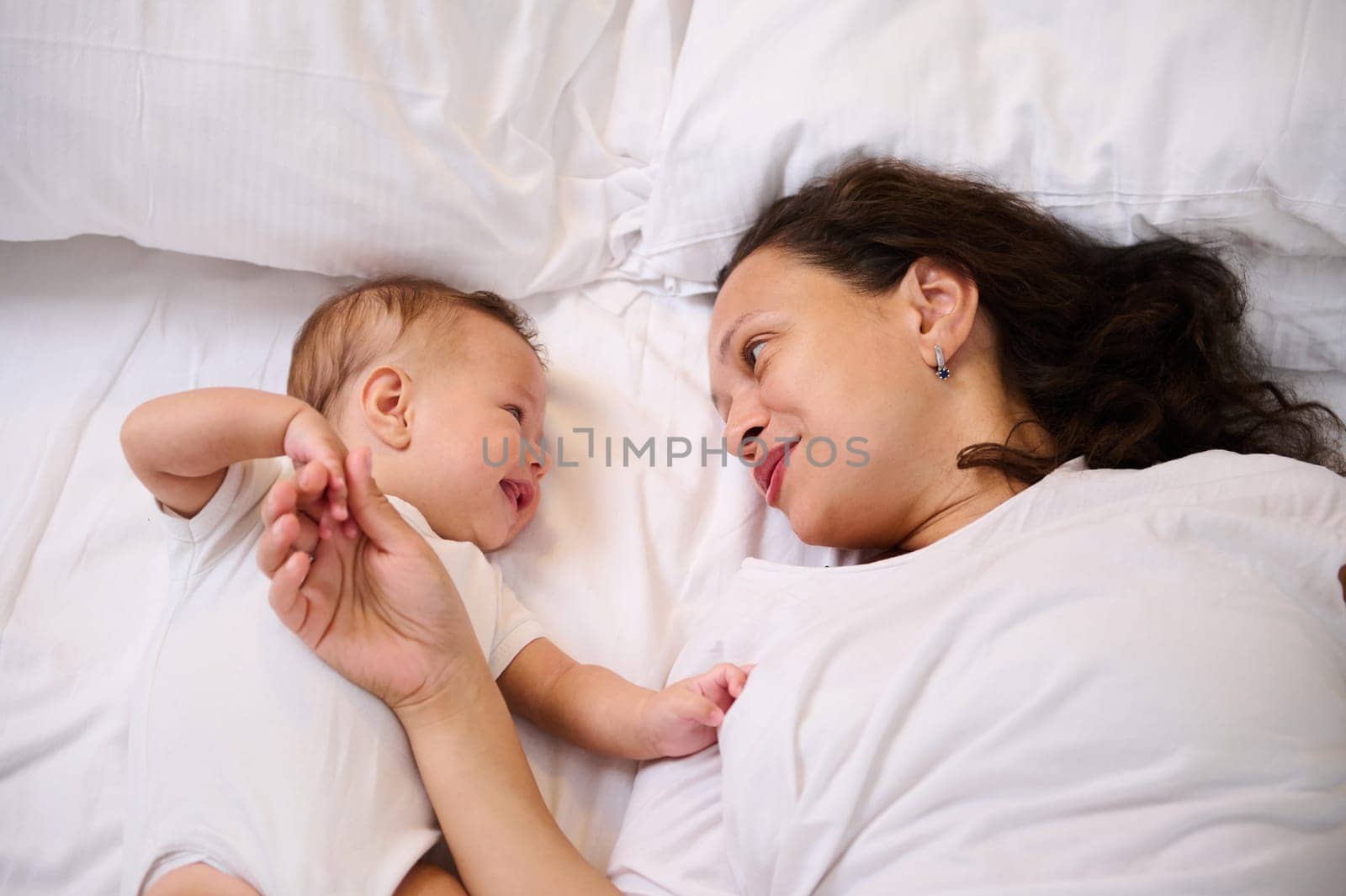 Adorable little baby have fun with his mom, smiling to her while lying together on the bed in the morning. Babyhood and maternity leave concept. Mother and child together