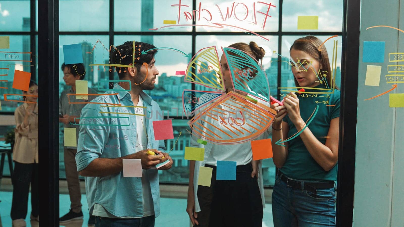 Group of happy professional business team working together, discussing and writing at sticky notes about marketing idea, plan, strategy at glass wall with graph and colorful sticky notes. Tracery
