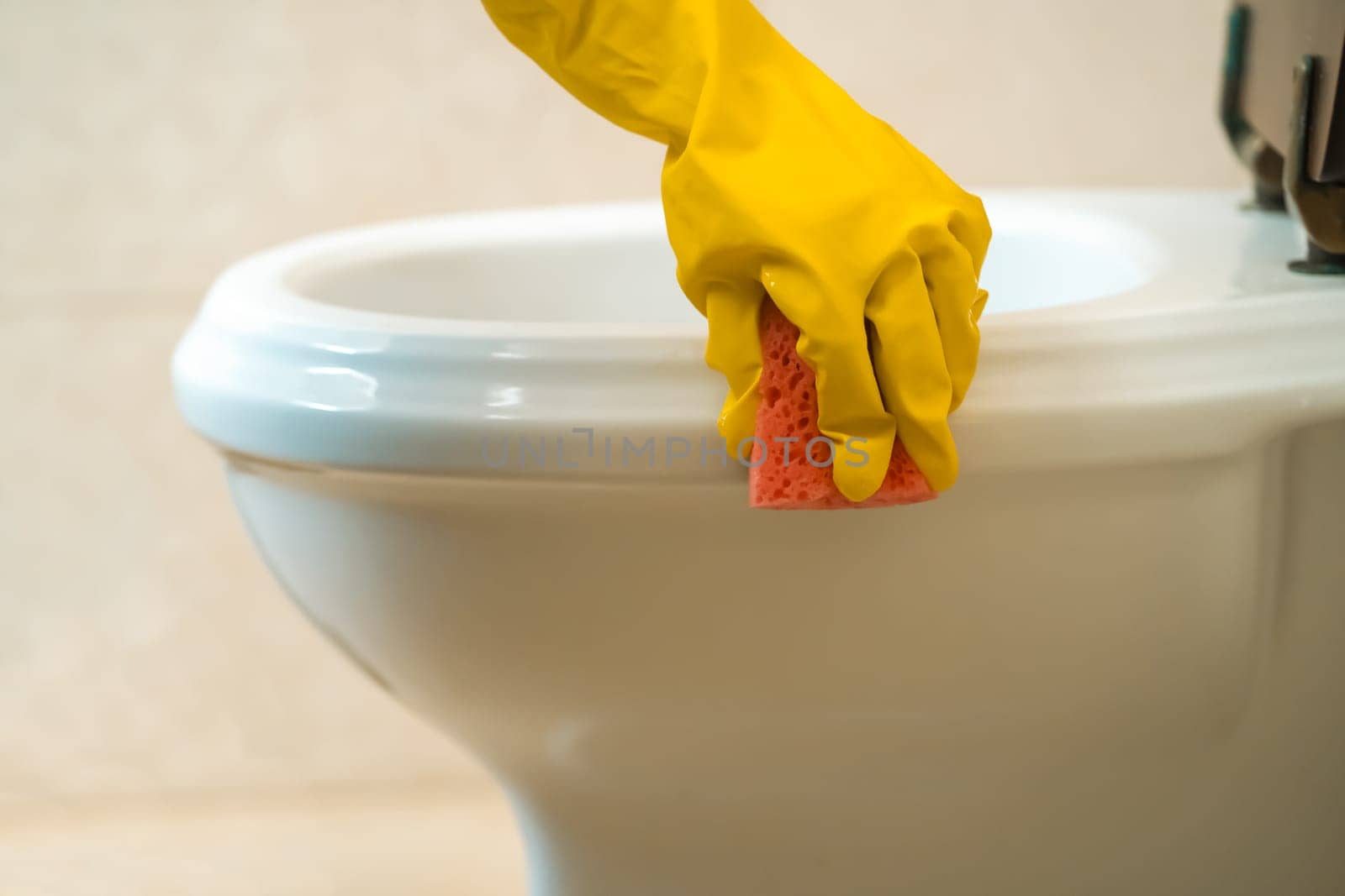 Female hands in yellow rubber gloves are holding a rag and wiping the toilet with detergent and disinfectant. The woman is cleaning the bathroom.