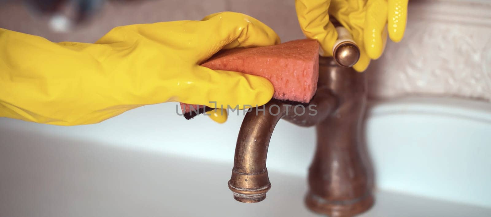 A woman in rubber gloves is cleaning the house, creating cleanliness and disinfection, wiping the faucet and wash basin in the bathroom with a sponge.