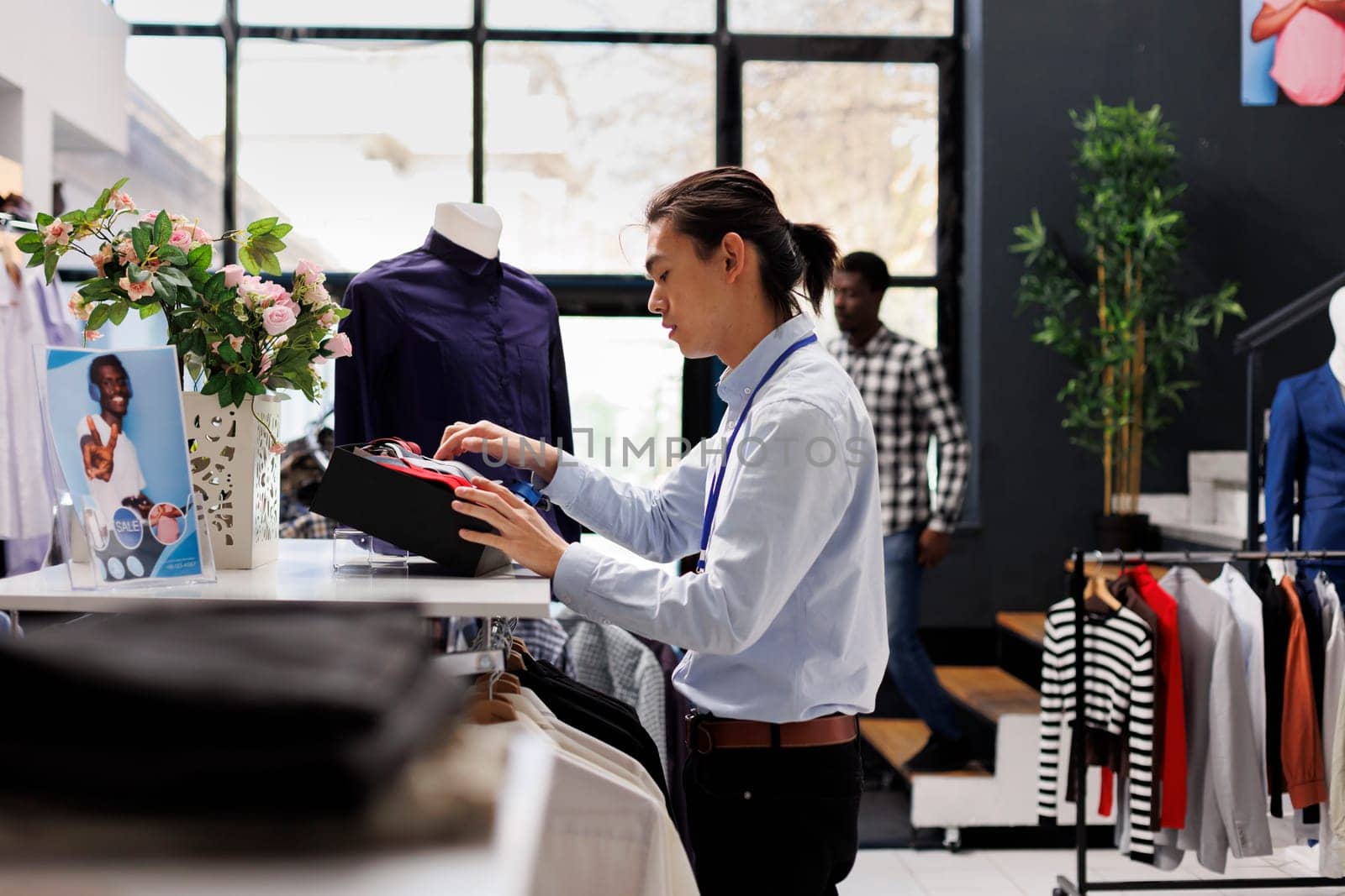 Asian manager arranging elegant tie and clothes, preparing clothing store for opening. Employee working with fashionable merchandise and accessories, checking new fashion collection in modern boutique