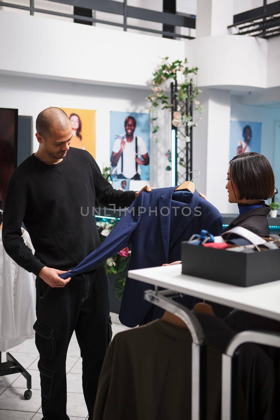 Arab man examining apparel size and style while talking with clothing store consultant. Shopping center fashion department employee giving buyer advice in selecting formal jacket