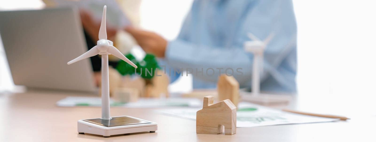 Windmill model represented using clean energy and wooden block represented eco house was scatter around on the table in front of businessman working on laptop. Blurring background. Delineation.