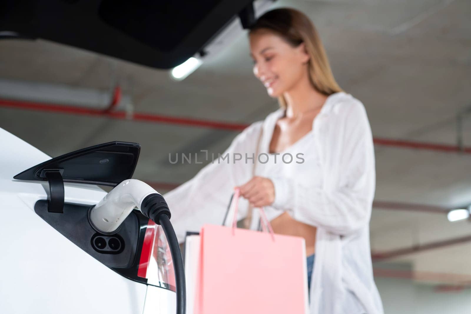 Young woman travel with EV electric car to shopping center parking lot charging in downtown city showing urban sustainability lifestyle by green clean rechargeable energy of electric vehicle innards