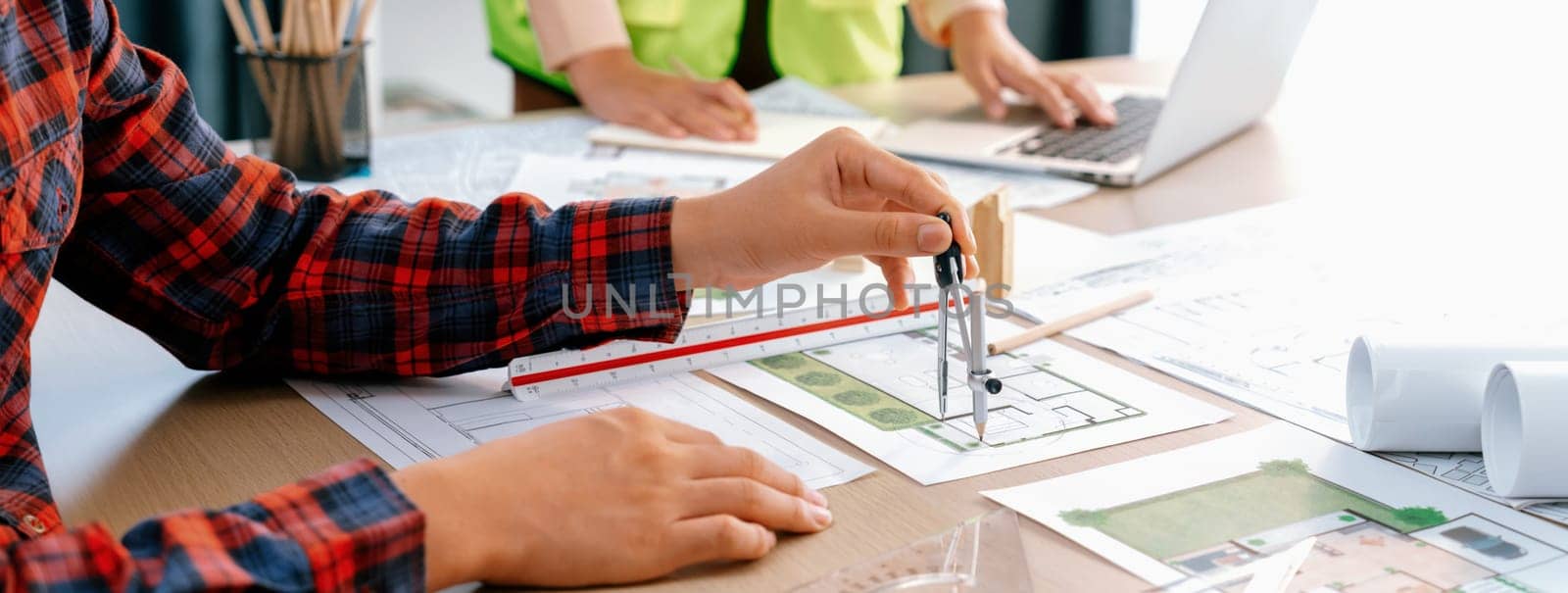 Cropped image of professional engineer team working on blueprint while his coworker working on laptop at meeting table with blueprint and wooden block scattered around. Closeup. Delineation.