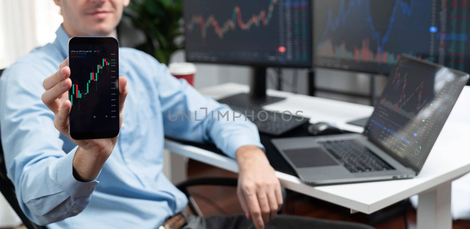Successful young business trader showing dynamic stock market graph on smartphone, presenting infomation database of currency exchange rate in real time against on pc blurred background. Gusher.