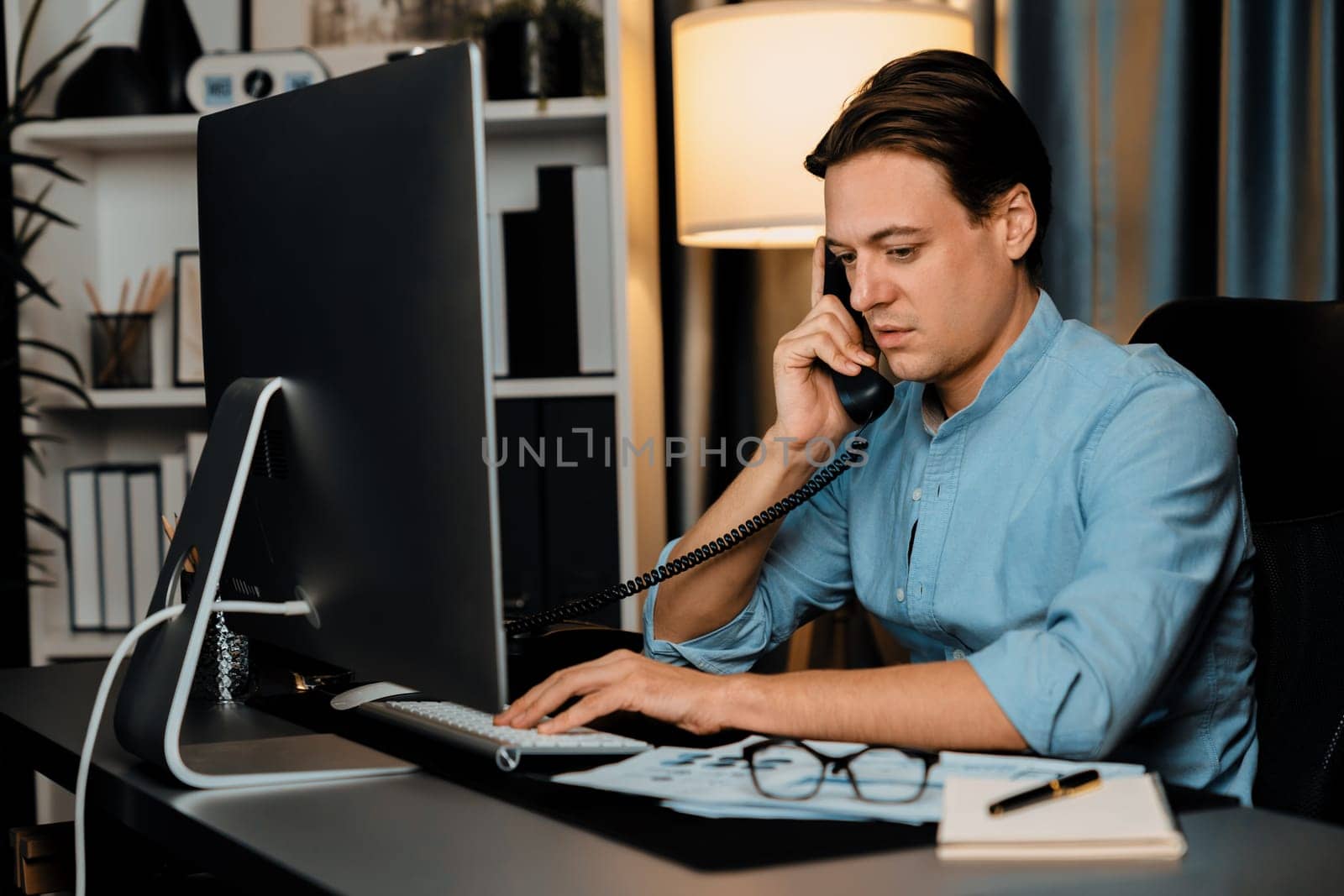 Smart businessman calling with telephone office for consulting business paragraph dynamic data marketing analysis plan surrounded paper sheet and pc monitor report at home office at night. Pecuniary.