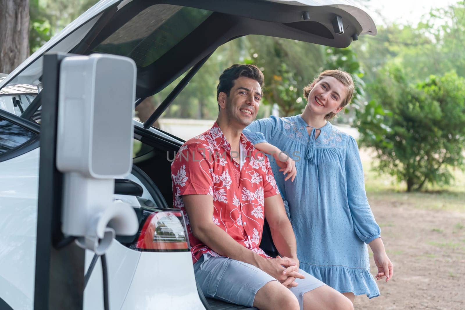 Road trip vacation with electric vehicle, lovely couple sitting on trunk and recharge EV car with green and clean energy. Nature and travel with eco-friendly car for sustainable environment. Perpetual