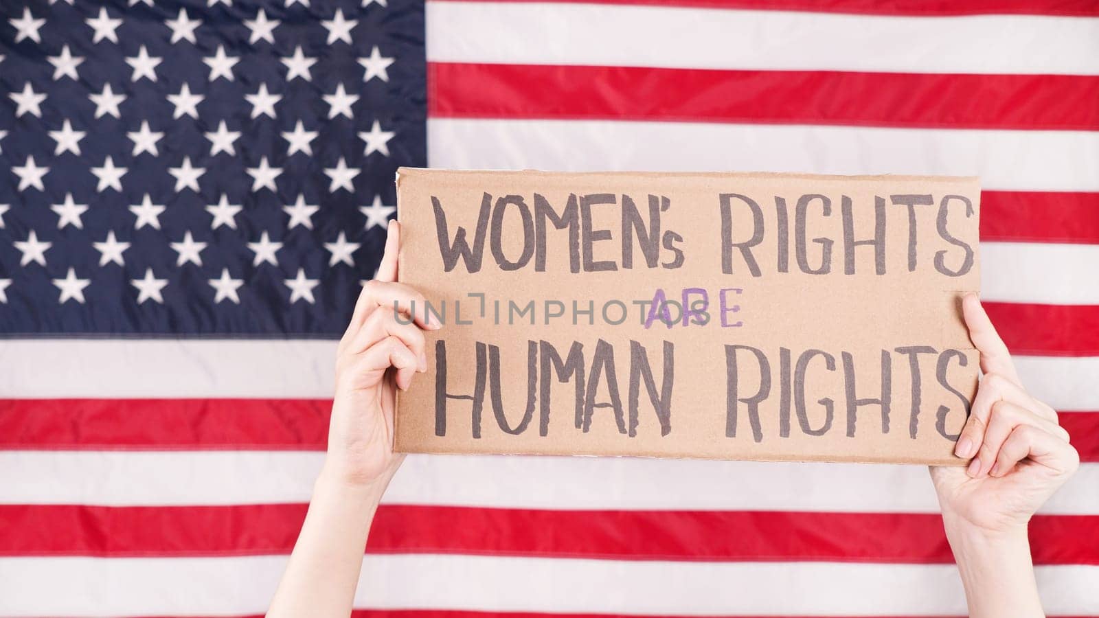 Young woman protester holds cardboard with Womens Rights Are Human Rights sign against USA flag on background. Feminist power. Equal opportunity Womens rights freedom. by JuliaDorian