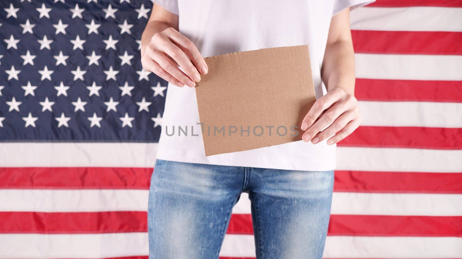 Unrecognizable woman showing empty cardboard banner. Hands holding blank poster from a cardboard box. Space for your slogan, logo or advertisement. Banner design concept. American flag on background by JuliaDorian