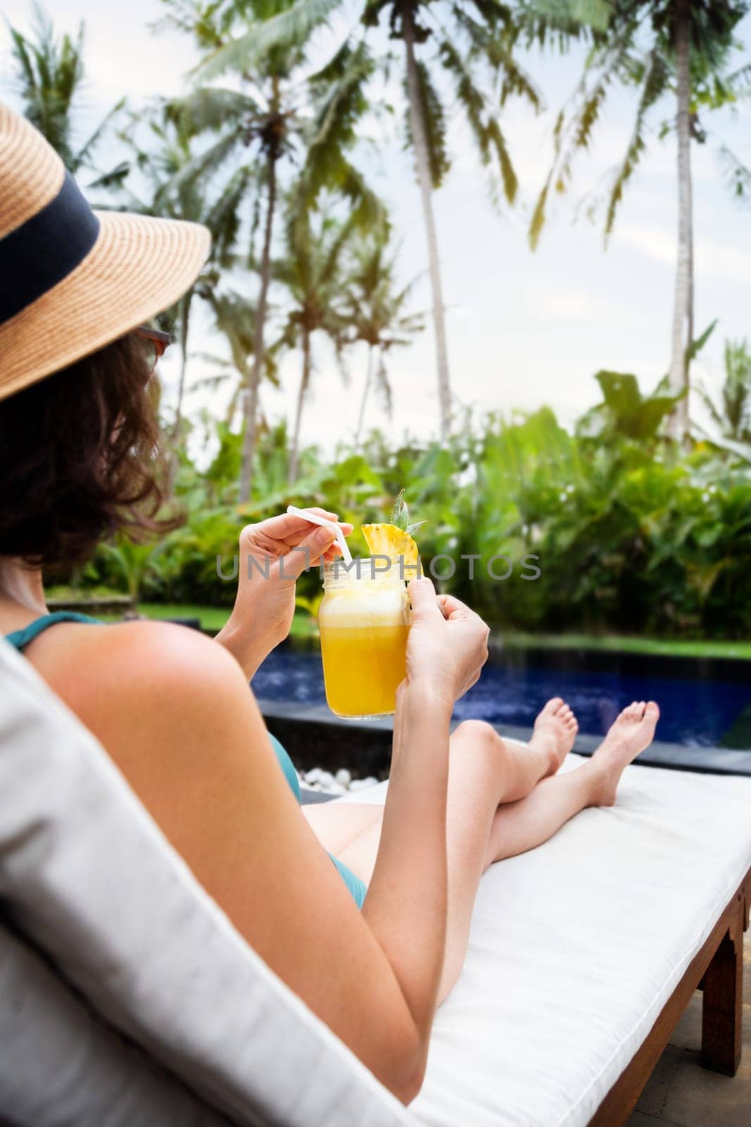 Woman relaxing sitting in desk chair near swimming pool drinking healthy pineapple juice during summer holidays.Vertical by Hoverstock