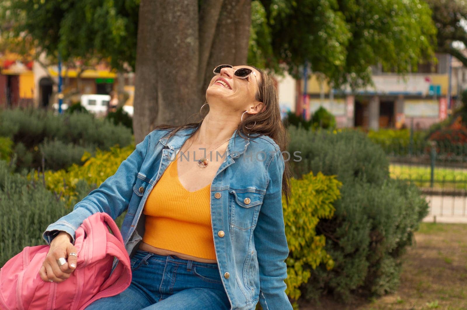 A woman, wearing jeans and a denim jacket, sits under a tree in the park, appearing cool and relaxed.
