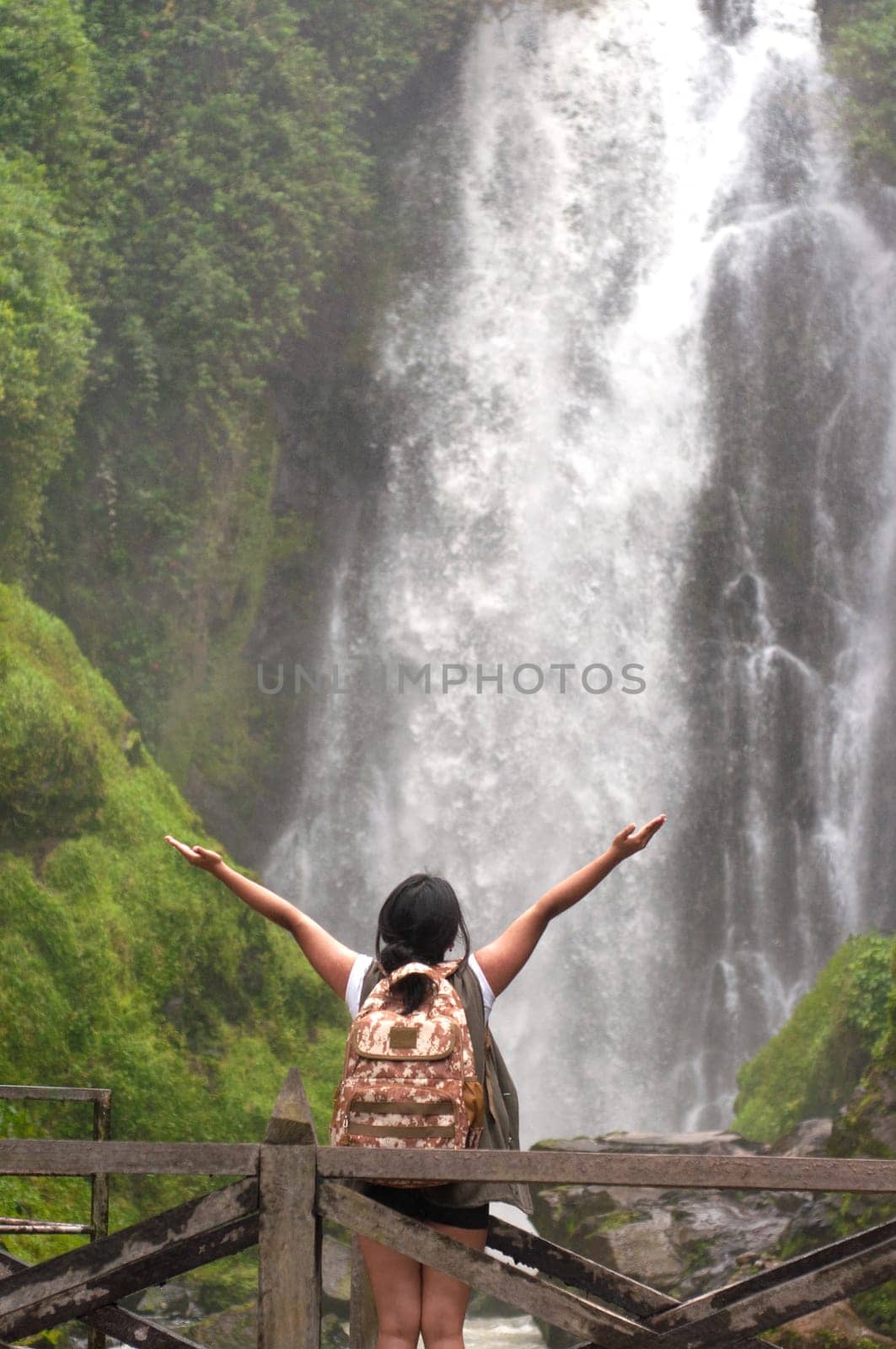 A lone traveler enjoys a moment of tranquility as she stands with arms outstretched facing a powerful waterfall surrounded by vibrant green foliage. by Raulmartin