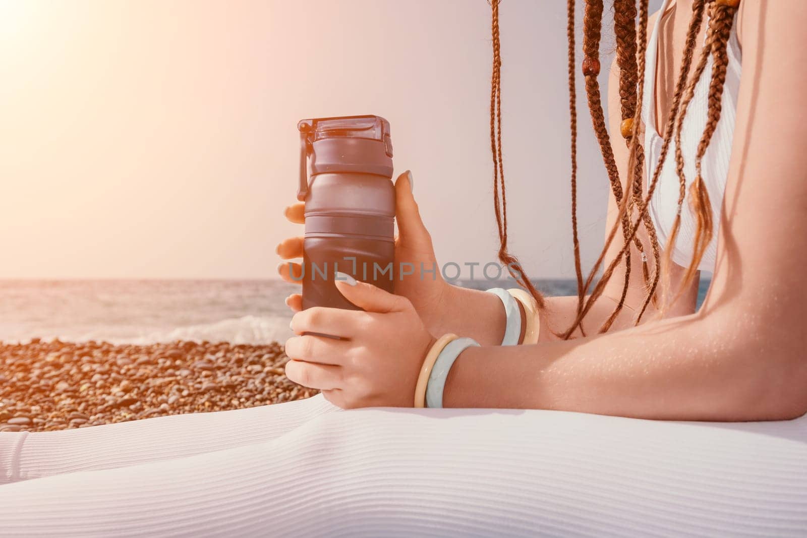 Woman sea yoga. Thermo mug in the hands of woman with braids dreadlocks in white leggings and tops doing stretching pilates on yoga mat near sea. Female fitness yoga routine concept. Healthy lifestyle by panophotograph