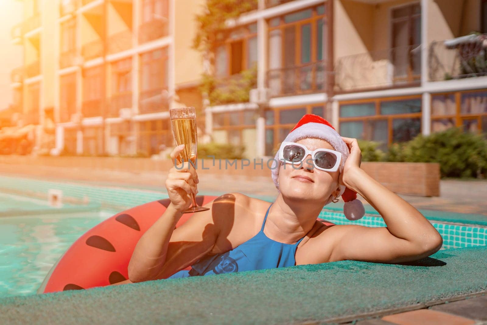 Woman pool Santa hat. A happy woman in a blue bikini, a red and white Santa hat and sunglasses poses near the pool with a glass of champagne standing nearby. Christmas holidays concept