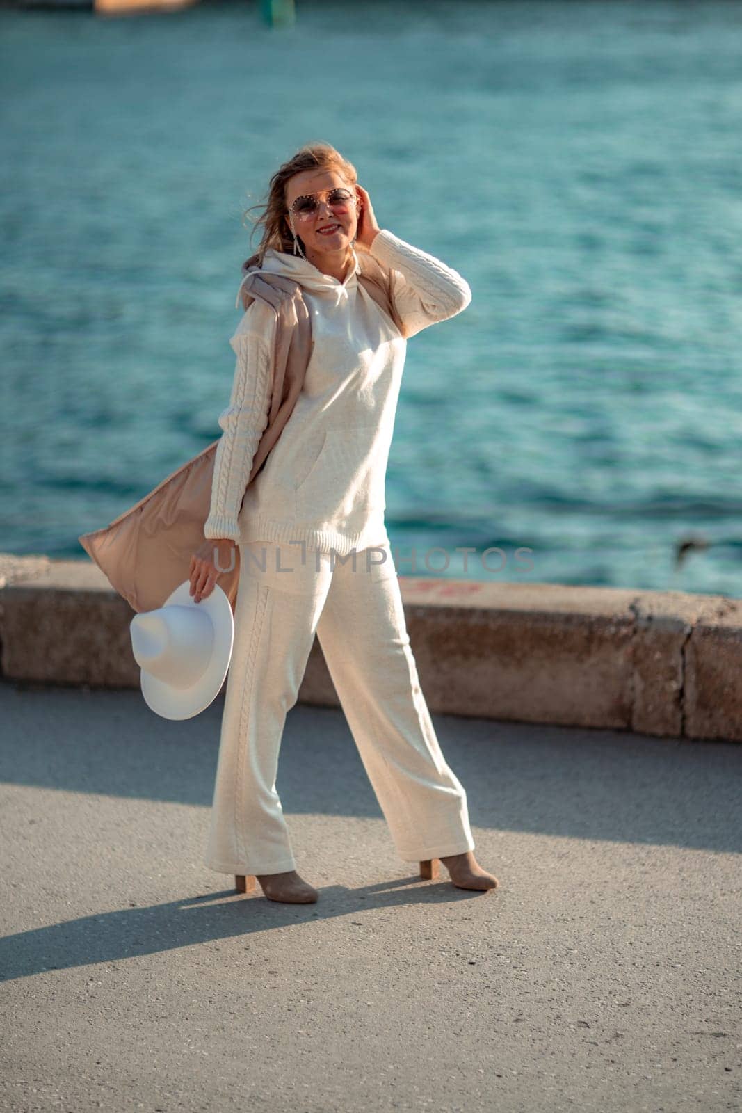 Happy blonde woman in a white suit and hat posing at the camera against the backdrop of the sea.