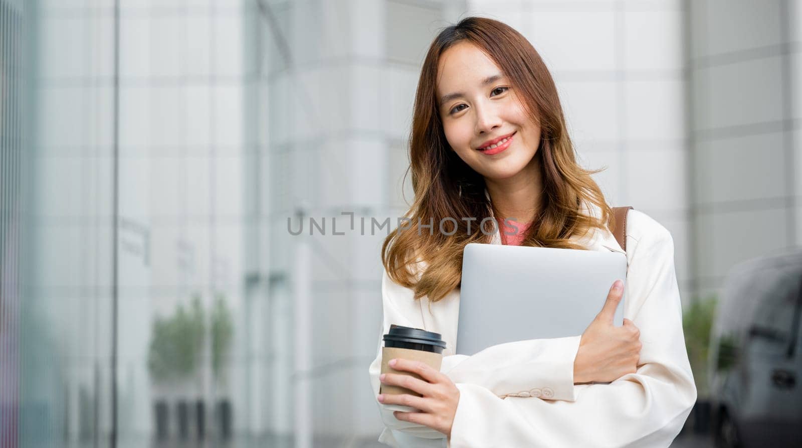 Smiling brunette girl walks down the street, carrying a paper cup of hot coffee and a laptop in her hands. city wall serves as a beautiful background for this portrait of productivity and multitasking
