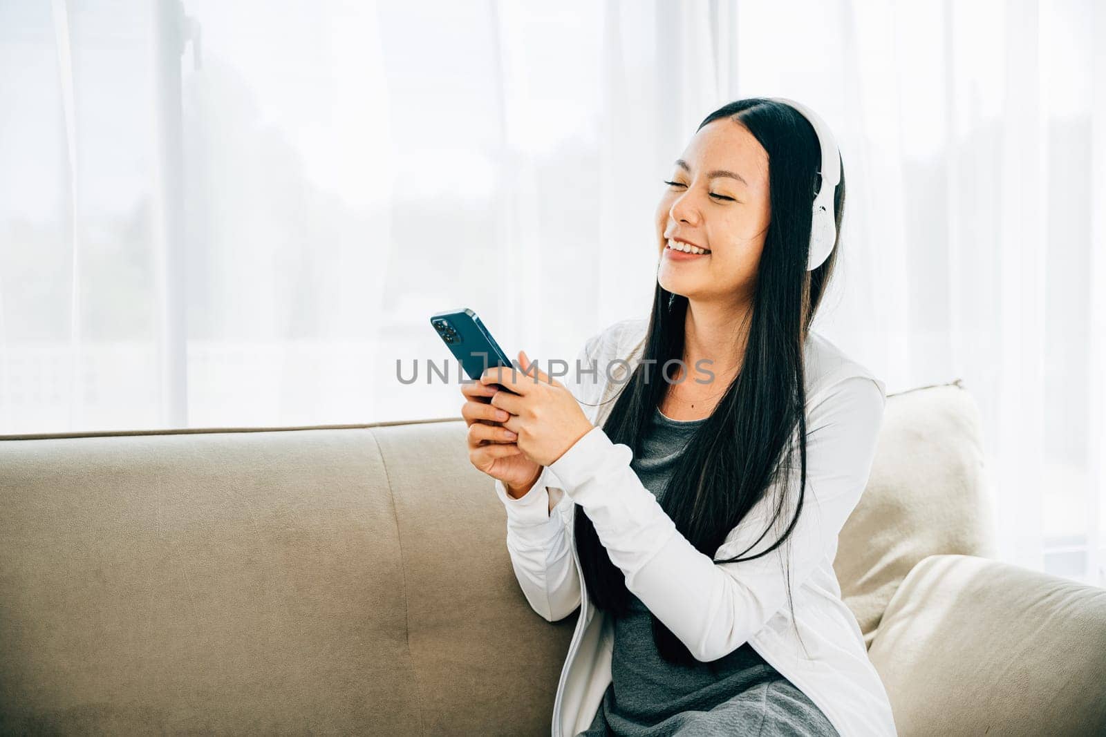 Woman with smartphone and headphones relaxes on sofa listens to music by Sorapop