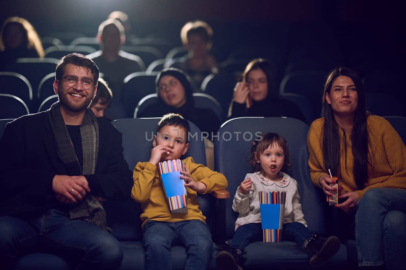 A modern family enjoys quality time together at the cinema, indulging in popcorn while watching a movie with their children.