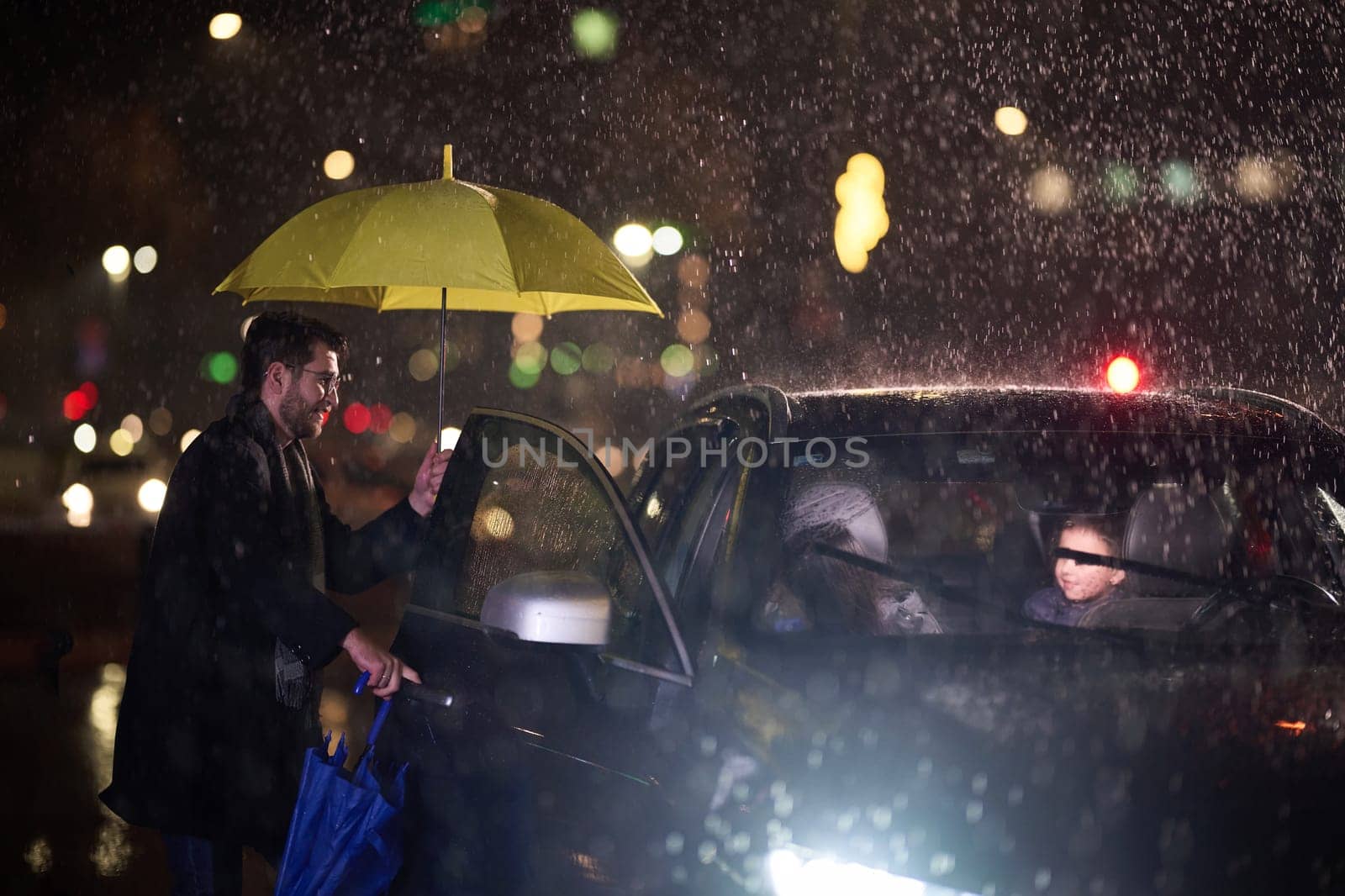 A family man lovingly opens the car for his family in rainy weather, sheltering them with an umbrella.