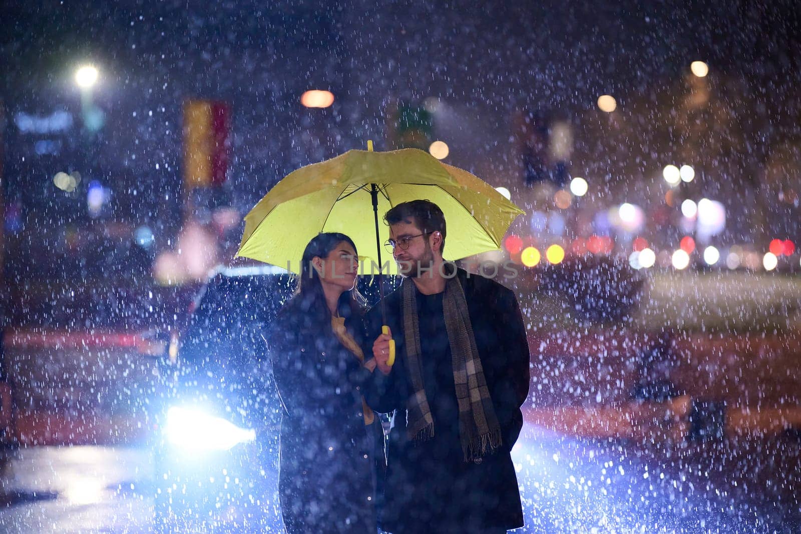 In the romantic ambiance of a rainy night, a happy couple walks through the city, sharing tender moments under a yellow umbrella, surrounded by the glistening glow of urban lights.